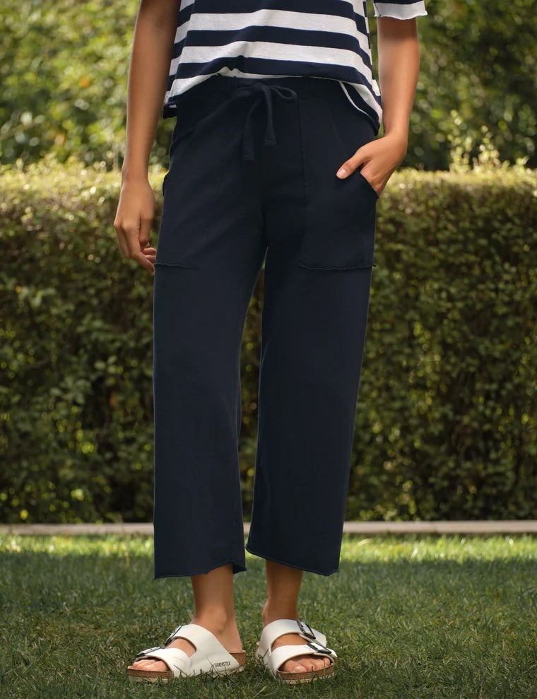 A person wearing a striped shirt, white sandals, and the FRANK & EILEEN PENNY Patch Pocket Jersey Pant in British Royal Navy stands on grass with a hedge and greenery in the background.