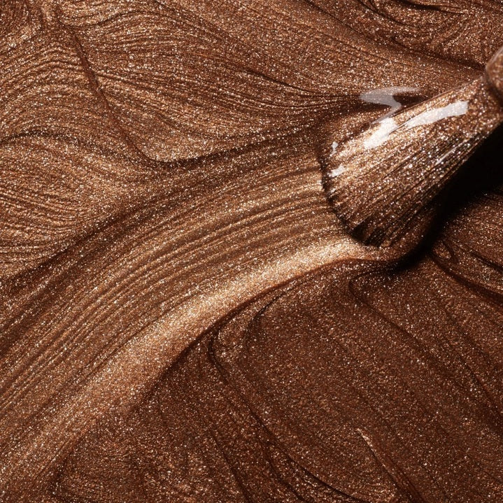A hand with shimmery bronze vegan, cruelty-free nail polish holds a matching bottle labeled “MANUCURIST - GREEN™ Nail Polish in Tanned” against a brown sweater.
