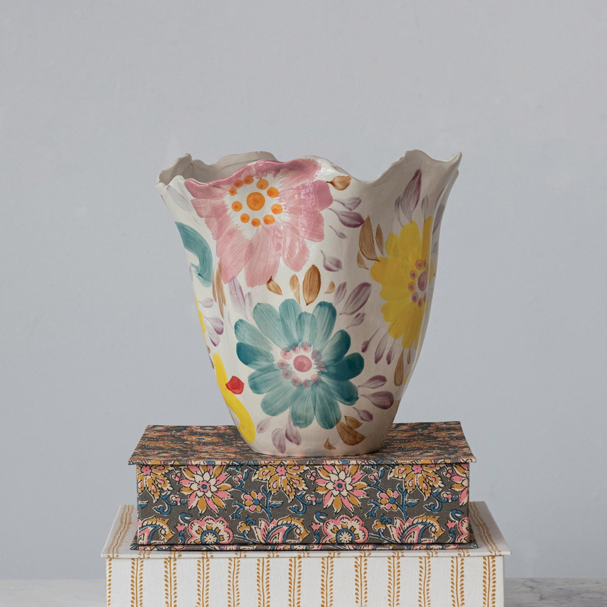 The RUFFLED VASE W FLORALS by CREATIVE COOP, crafted from stoneware materials and featuring a scalloped rim, is displayed atop a pair of decorative books. The vase is beautifully adorned with large flowers in vibrant shades of blue, yellow, pink, and orange. The ornate books have eye-catching covers—one flaunts a floral design while the other showcases vertical stripes.