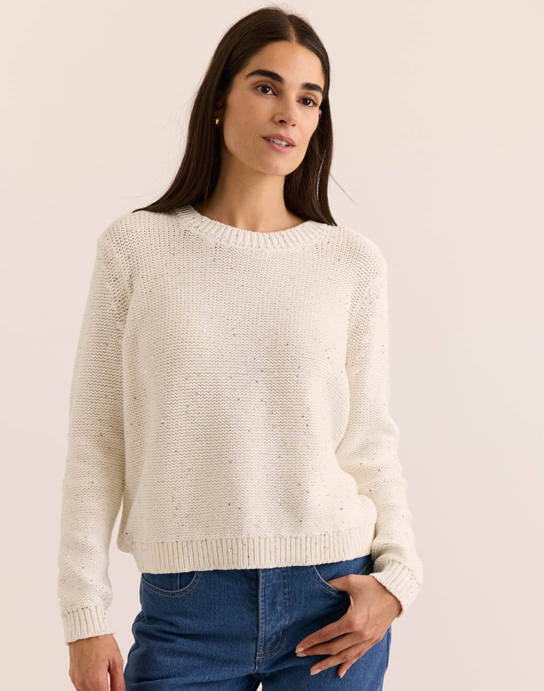 A woman with long brown hair wears the HINSON WU - QUEENIE Long Sleeve Sequin Sweater in Pearl White and blue jeans, standing against a plain light background and looking slightly to the side with a relaxed expression.