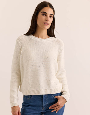 A woman with long brown hair wears the HINSON WU - QUEENIE Long Sleeve Sequin Sweater in Pearl White and blue jeans, standing against a plain light background and looking slightly to the side with a relaxed expression.