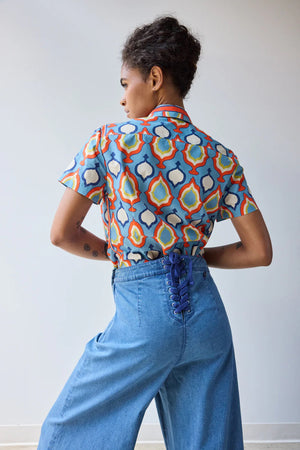 A person with short curly hair and tattoos wears the HOPE FOR FLOWERS SCARF TIE BUTTON DOWN SHIRT IN CHAMBRAY MEDALLION, tucked into high-waisted blue wide-leg jeans, posing against a plain light background.