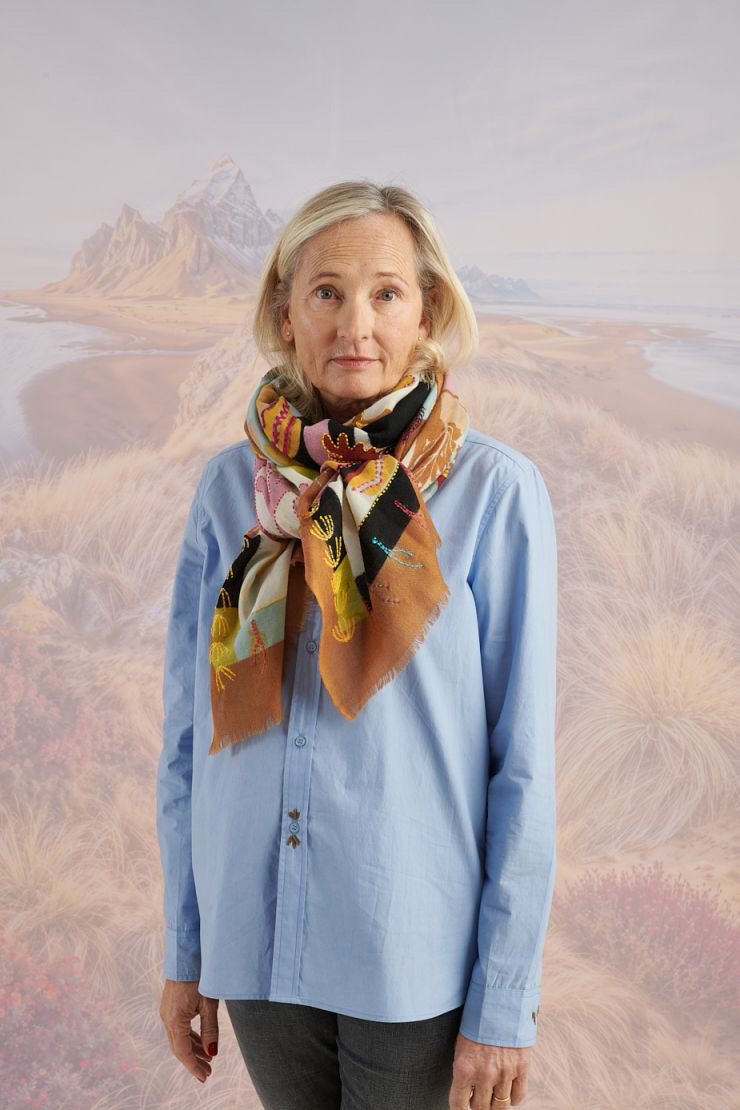A woman with light brown hair wears a cream blouse and the STORIATIPIC - ANDRES WOOL SCARF by STORIATIPIC, elegantly hand-embroidered with green, yellow, red, and blue patterns. She gazes to the side against a scenic background.
