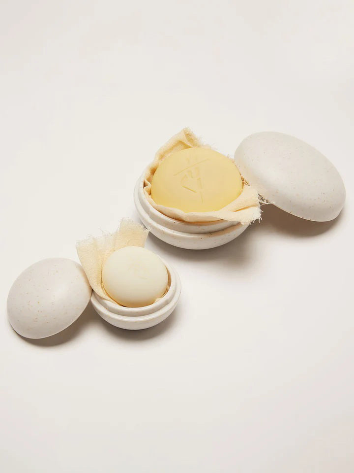 A round, creamy white lotion bar from KATE MCLEOD rests on a textured beige cloth in an open, speckled container. The lid beside it resembles a mini body stone. This minimalistic setup of THE MINI TRAVEL CASE by KATE MCLEOD is perfect for travel against a plain background.