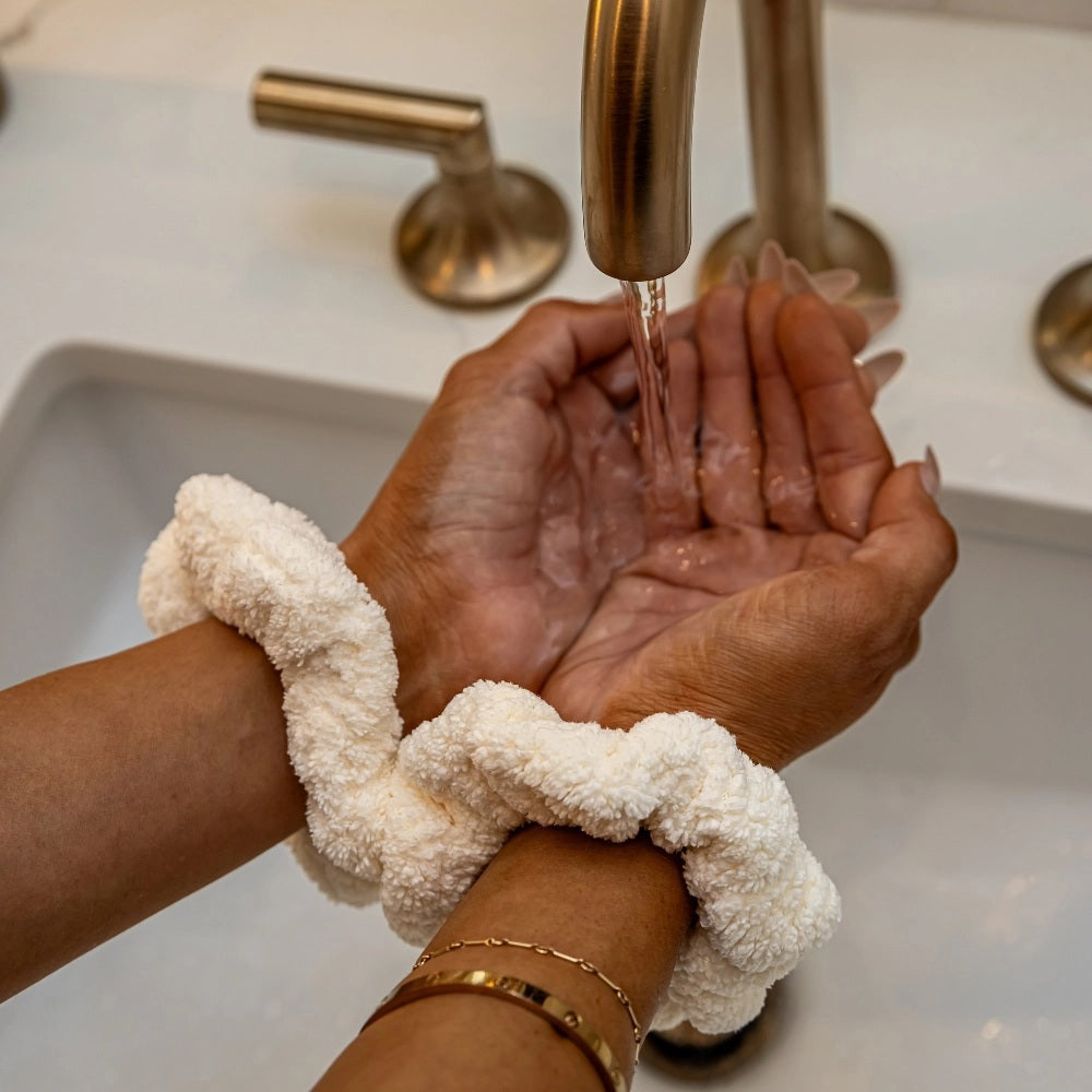 A woman with wavy, light brown hair in a ponytail secured with a VOLO BEAUTY - SCRUNCHIE DUO PACK is shown from the back, wearing a black shirt and gold jewelry, and raising her left hand.