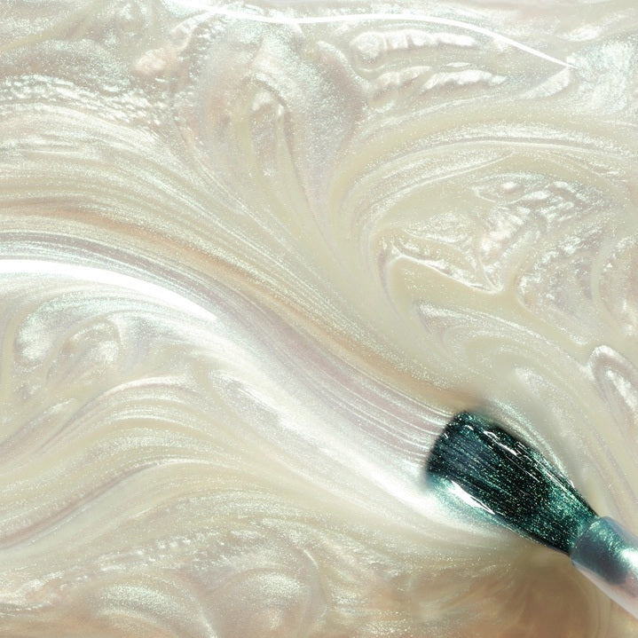 A person with light green painted nails holds a bottle of MANUCURIST INC’s GREEN™ Nail Polish in Fairy, wearing a purple knit top adorned with rhinestone embellishments.