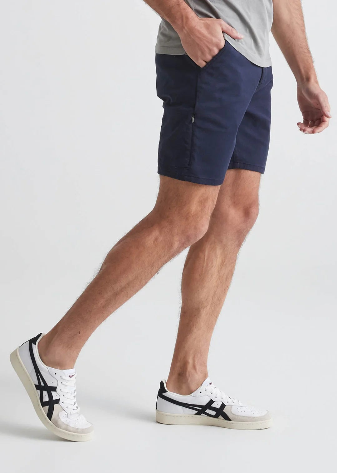 A man in a light blue T-shirt, DUER Live Free Journey Shorts in Sapphire, and white sneakers with black details stands with one hand in his pocket against a plain light background.