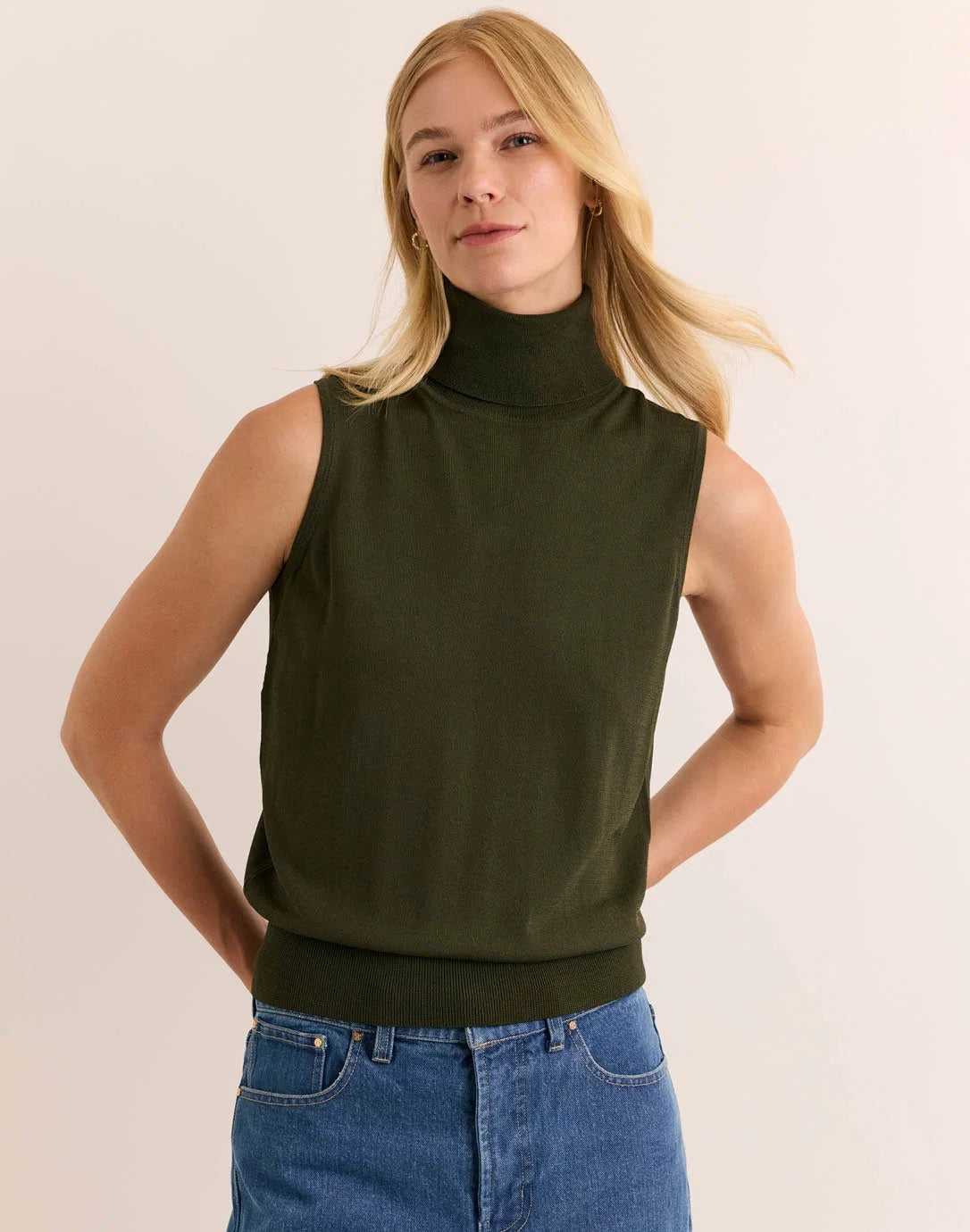 A woman with long blonde hair wears the HINSON WU - ASHLEY Sleeveless Turtleneck Knit Tank in Oregano, crafted from lightweight Verona Knit yarn, paired with blue jeans. She stands against a plain light background with one hand resting on her hip.