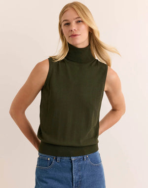 A woman with long blonde hair wears the HINSON WU - ASHLEY Sleeveless Turtleneck Knit Tank in Oregano, crafted from lightweight Verona Knit yarn, paired with blue jeans. She stands against a plain light background with one hand resting on her hip.