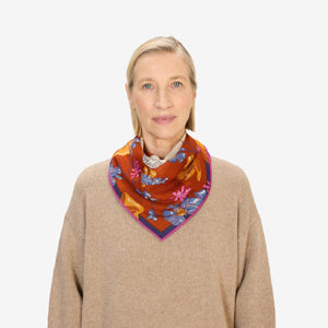 A woman with blonde hair, wearing a tan sweater and the INOUI TOUTOU DOG SCARF IN ORANGE, stands facing the camera against a plain white background.