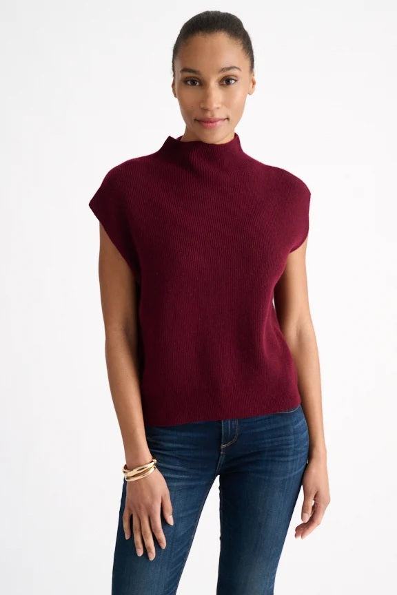 A woman is seen standing against a plain white background, smiling gently with one hand relaxed by her side, wearing the KINROSS CASHMERE Cap Sleeve Cashmere Rib Funnel in Cordovan from KINROSS, paired with blue jeans.