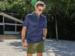 A man with a gray beard is wearing the CRIQUET Throwback Players Shirt in Navy, featuring a classic fit with white trim on the collar and a front pocket, as he stands outdoors surrounded by greenery.