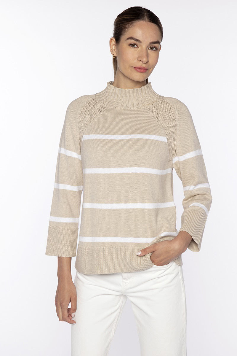 A woman with brown hair tied back wears the KINROSS STRIPE EASY FUNNEL NECK COTTON SWEATER in Dune/White and white pants, standing against a plain white background, smiling softly with one hand in her pocket.