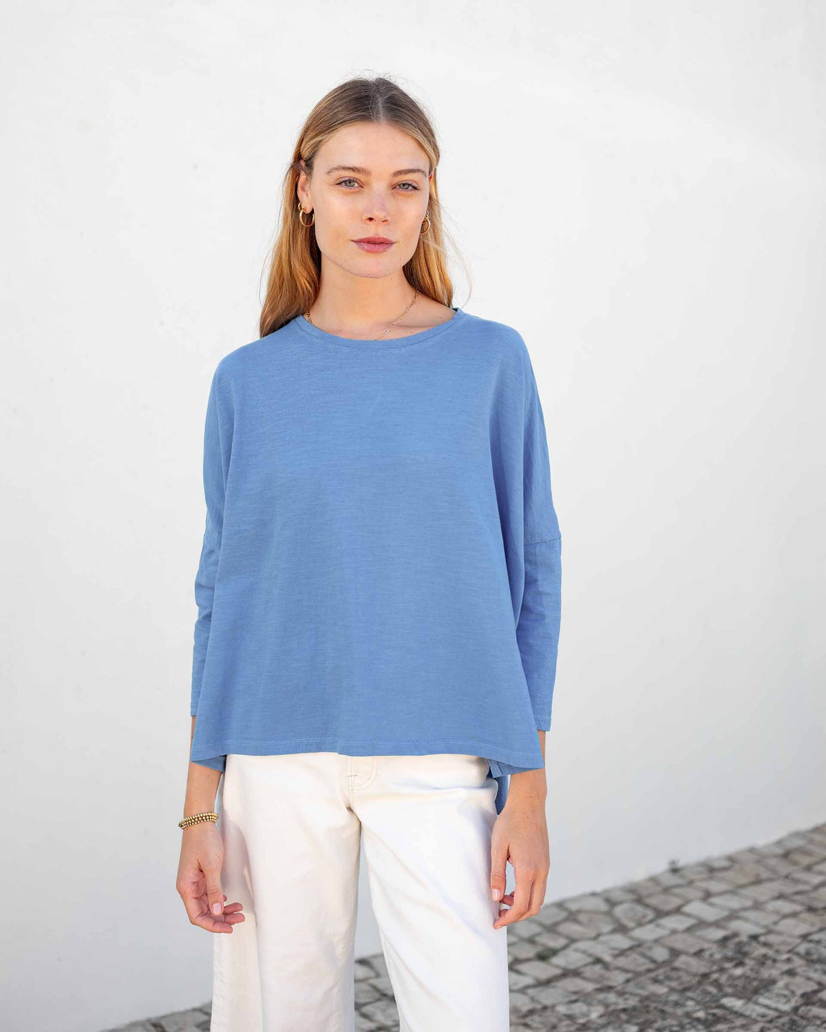 A woman with long, straight hair stands against a white wall, wearing the MERSEA Catalina Slub Tee in blue paired with white pants. The midweight textured cotton tee is from MERSEA. She looks at the camera with a neutral expression.