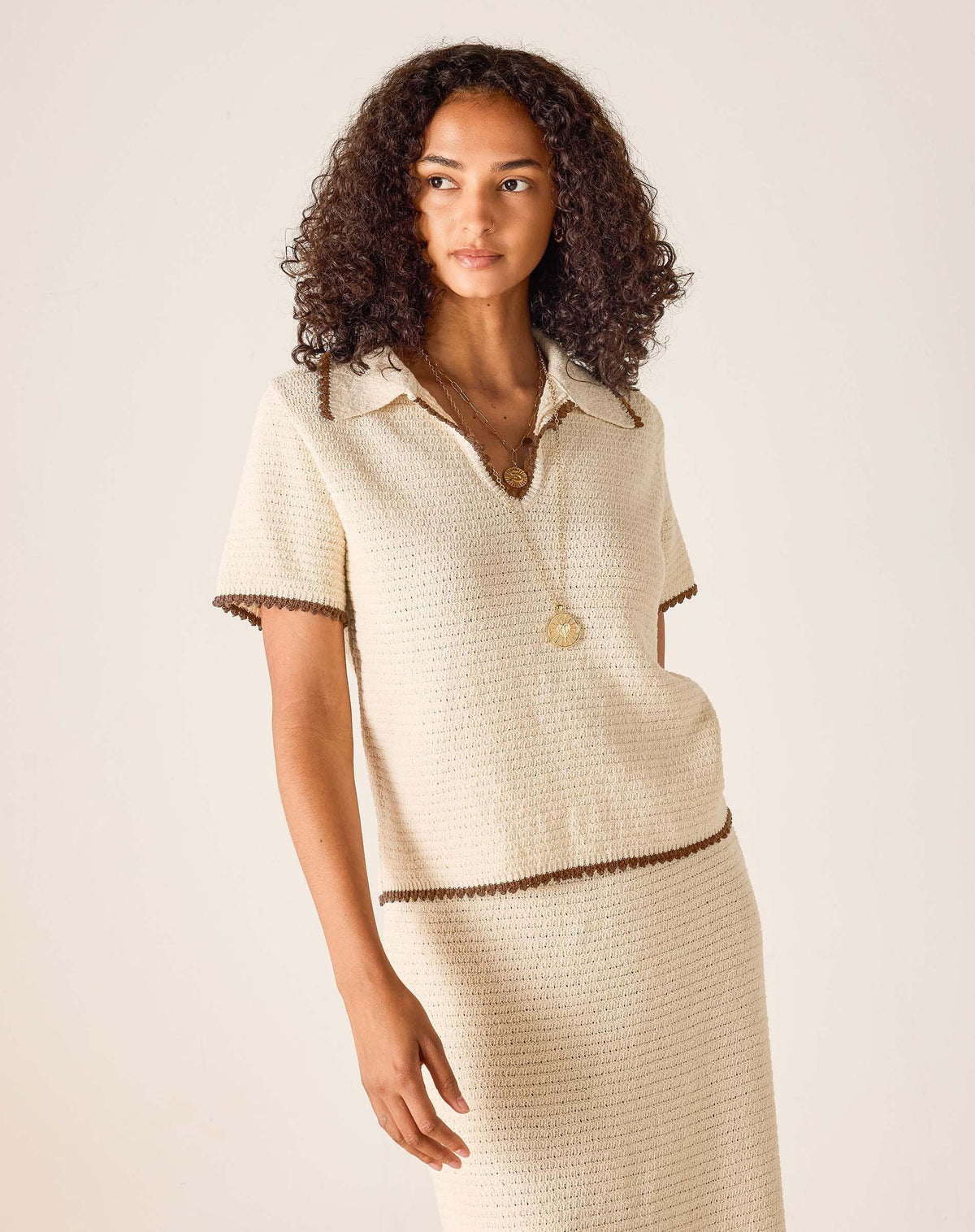 A woman stands in natural light near a window, wearing the MERSEA Paley Crochet Top in cream with brown trim and a matching skirt. Her wavy blonde hair is loose, and she poses against a white wall where various shadow patterns are visible.