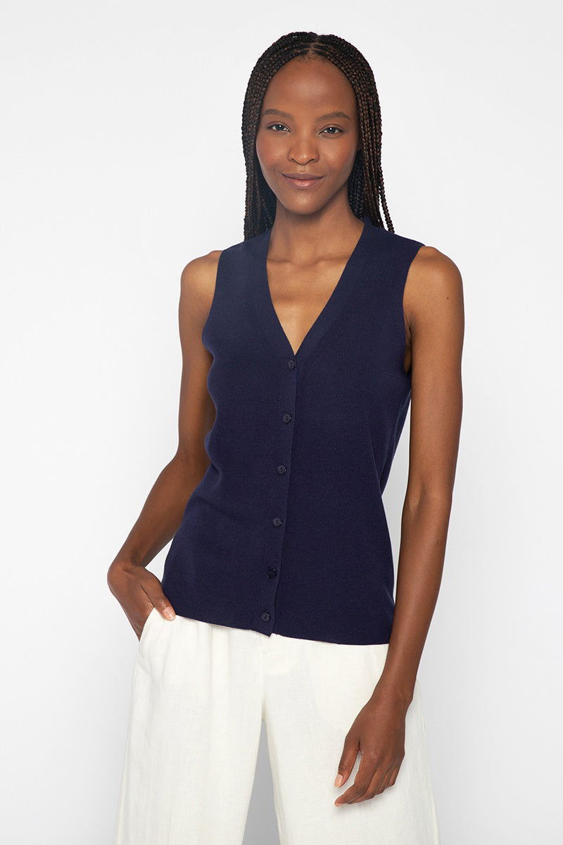 A woman with long braided hair is wearing the KINROSS CASHMERE - BUTTON VEST by KINROSS, a luxurious sleeveless navy blue button-up vest, paired with white pants. She is smiling and standing against a plain white background with one hand in her pocket.