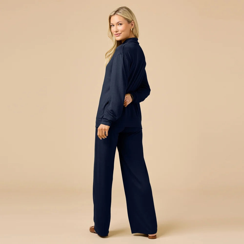 A woman wears a navy blue SOFTIES - DREAM JERSEY COWL NECK LOUNGE SET consisting of a long-sleeved top and wide-leg trousers.