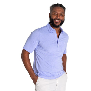 A smiling man with short curly hair and a beard wears the CRIQUET Top-Shelf Players Shirt in Bluebonnet Microstripe and light-colored pants, standing with his hands in his pockets against a white background.