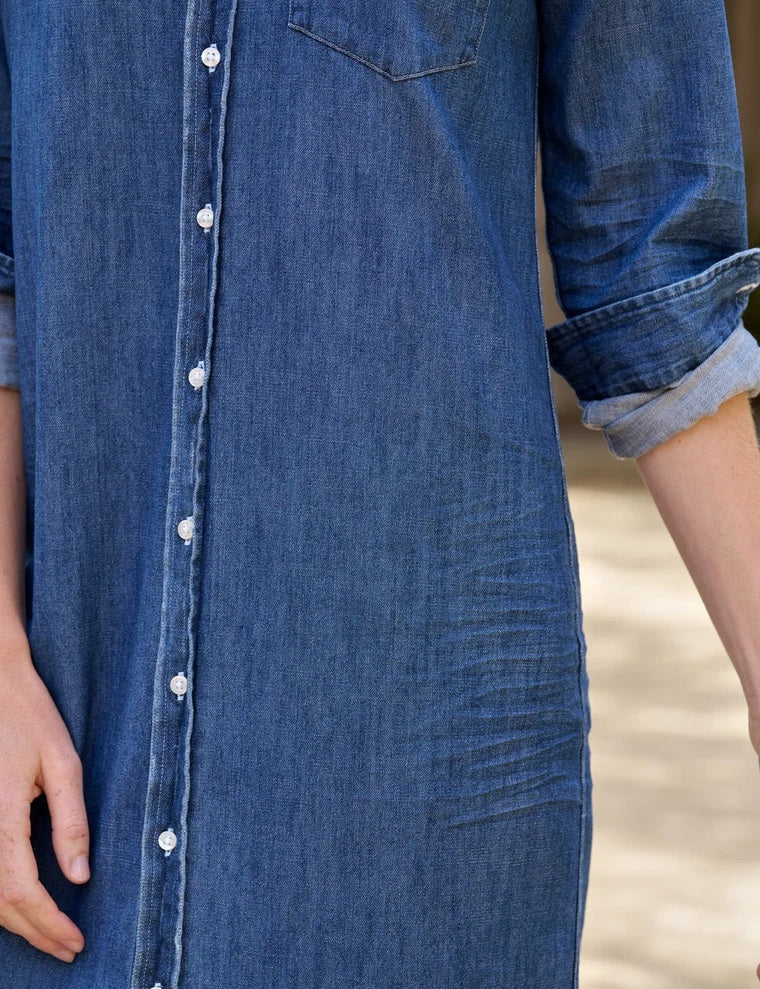 A woman stands outdoors in the FRANK & EILEEN Rory Maxi Shirtdress in Distressed Indigo Washed Denim, its long button-up silhouette and rolled sleeves crafted from luxurious indigo fabric. She pairs the effortlessly chic dress with brown knee-high boots, set against a backdrop of lush greenery and a building.