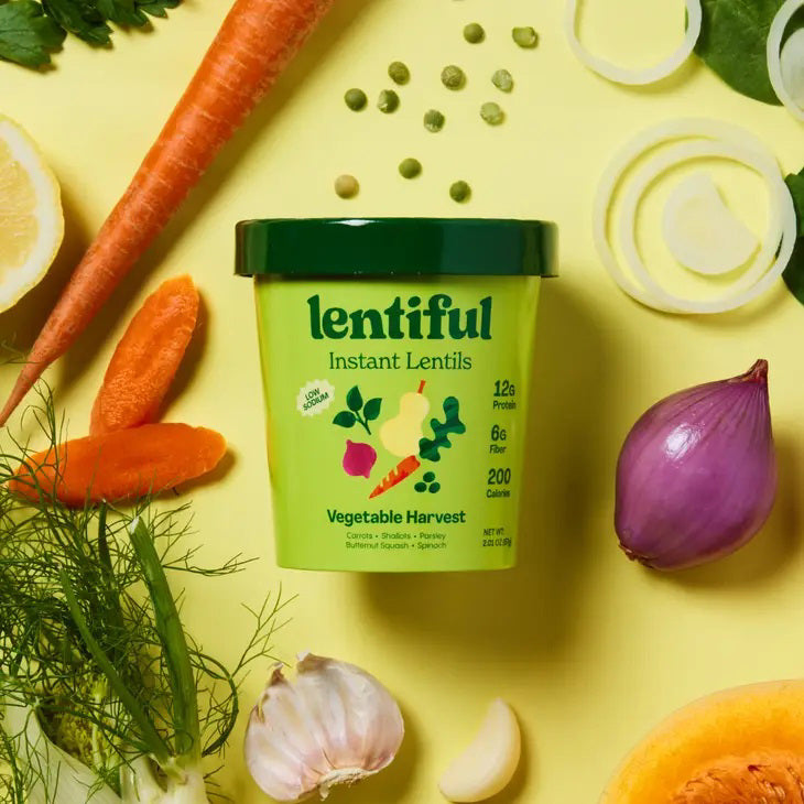 A container of LENTIFUL - LOW SODIUM VEGETABLE HARVEST INSTANT LENTILS by LENTIFUL sits on a yellow background, surrounded by fresh carrot, red onion, garlic, fennel, and squash.
