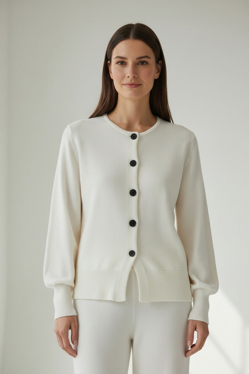 A woman stands indoors, facing the camera and smiling slightly against a plain light background, wearing the MEIMEIJ Button Up Jacket in Sugar Swizzle with a sculpted waist and matching white pants.