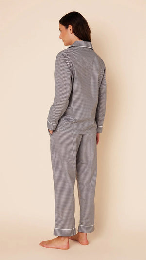A person with long dark hair is standing on a light beige background, wearing a matching set of loose-fitting, long-sleeved CAT'S PAJAMAS - CLASSIC GINGHAM LUXE PIMA PAJAMA by CAT'S PAJAMAS. Made from Luxe Pima Cotton, the buttoned-up pajamas feature a collar. The person is barefoot and looking slightly towards the camera.