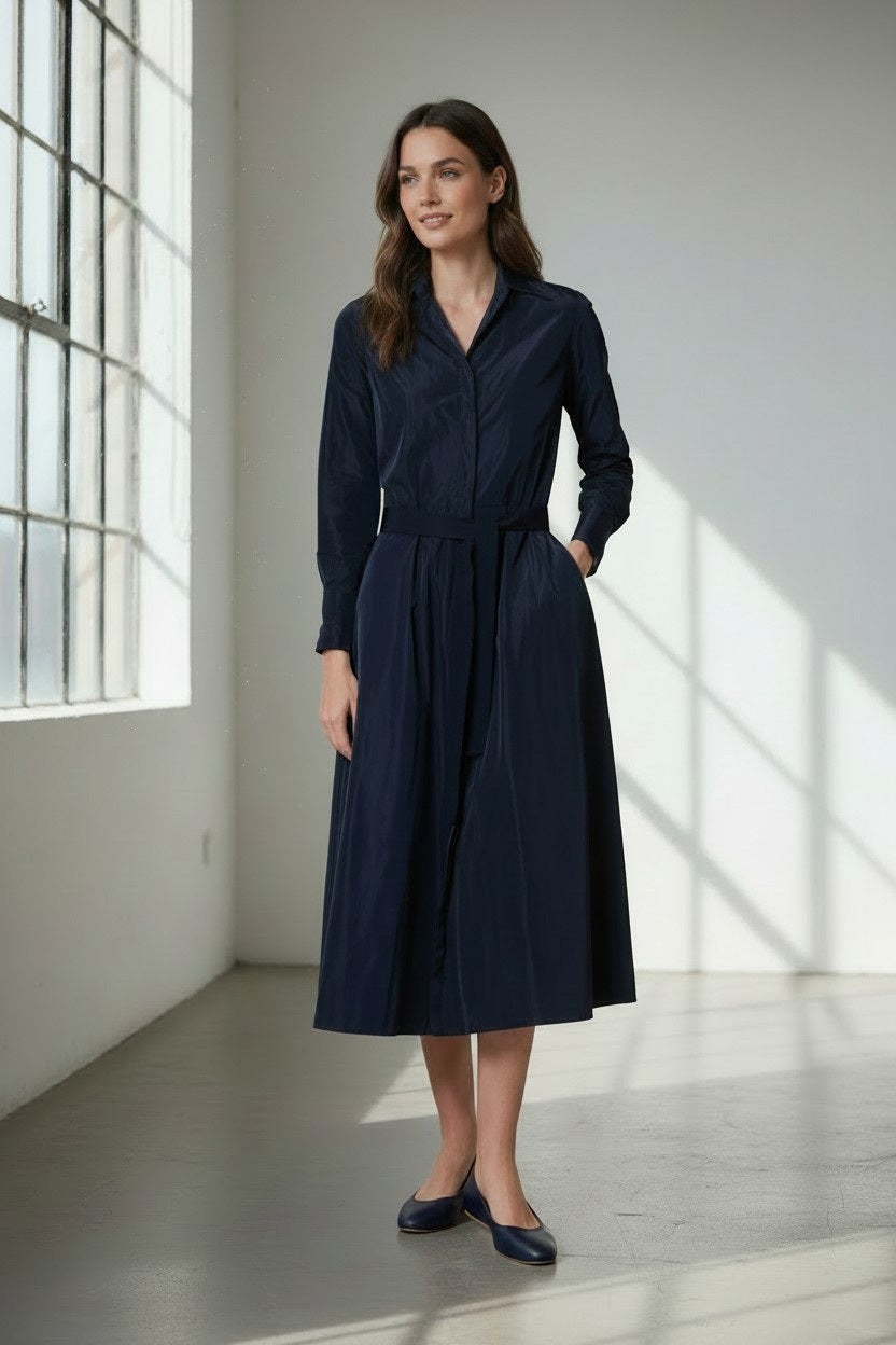 A woman stands in a sunlit room with her hands in her pockets and a slight smile, wearing the VICARIO CINQUE - ANNA Taffeta Midi Shirt Dress in midnight navy by VICARIO CINQUE, belted at the waist and paired with matching flats.
