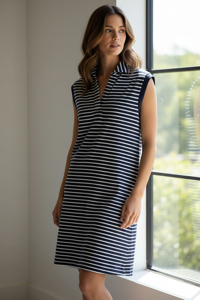 A person smiles outdoors, wearing the FRANK &amp; EILEEN Mini Lily Popover Henley Tank Dress in Vintage Jersey Yacht Stripe, a sleeveless navy and white striped dress made in California. Lush greenery and a bright blue sky appear in the background.