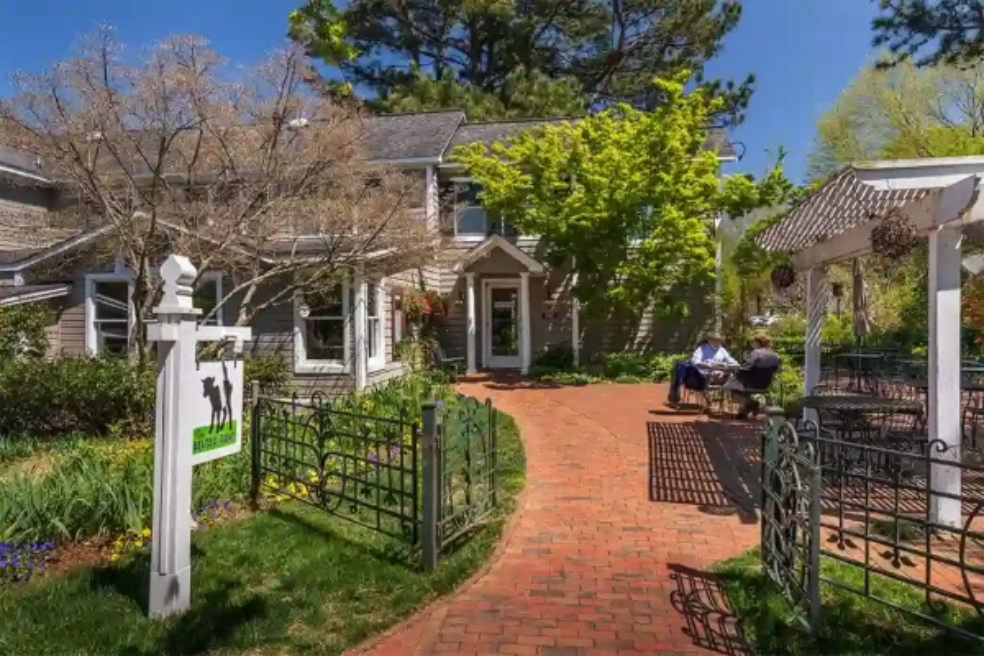 A brick pathway leads to a charming house with a white arbor and green garden. Two people are seated at a table under the arbor, and a real estate sign is visible on the left. Trees and plants surround the area.