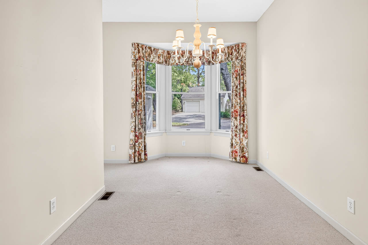 A bright, empty room with beige carpet, light walls, a chandelier, and a bay window with floral curtains, offering a view of trees and a garage outside.