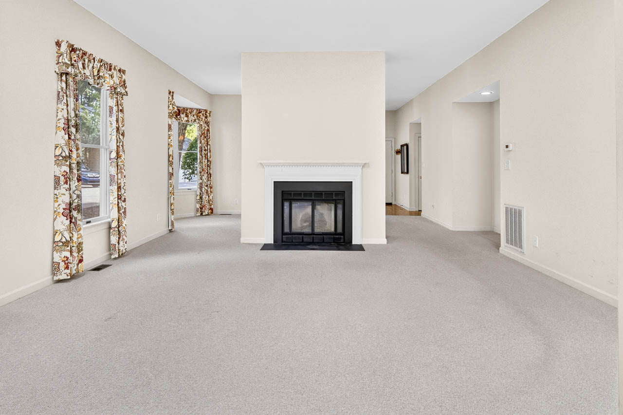 A spacious living room with beige walls and carpet, a central white fireplace, and two large windows with floral curtains letting in natural light. Hallways lead to other rooms.