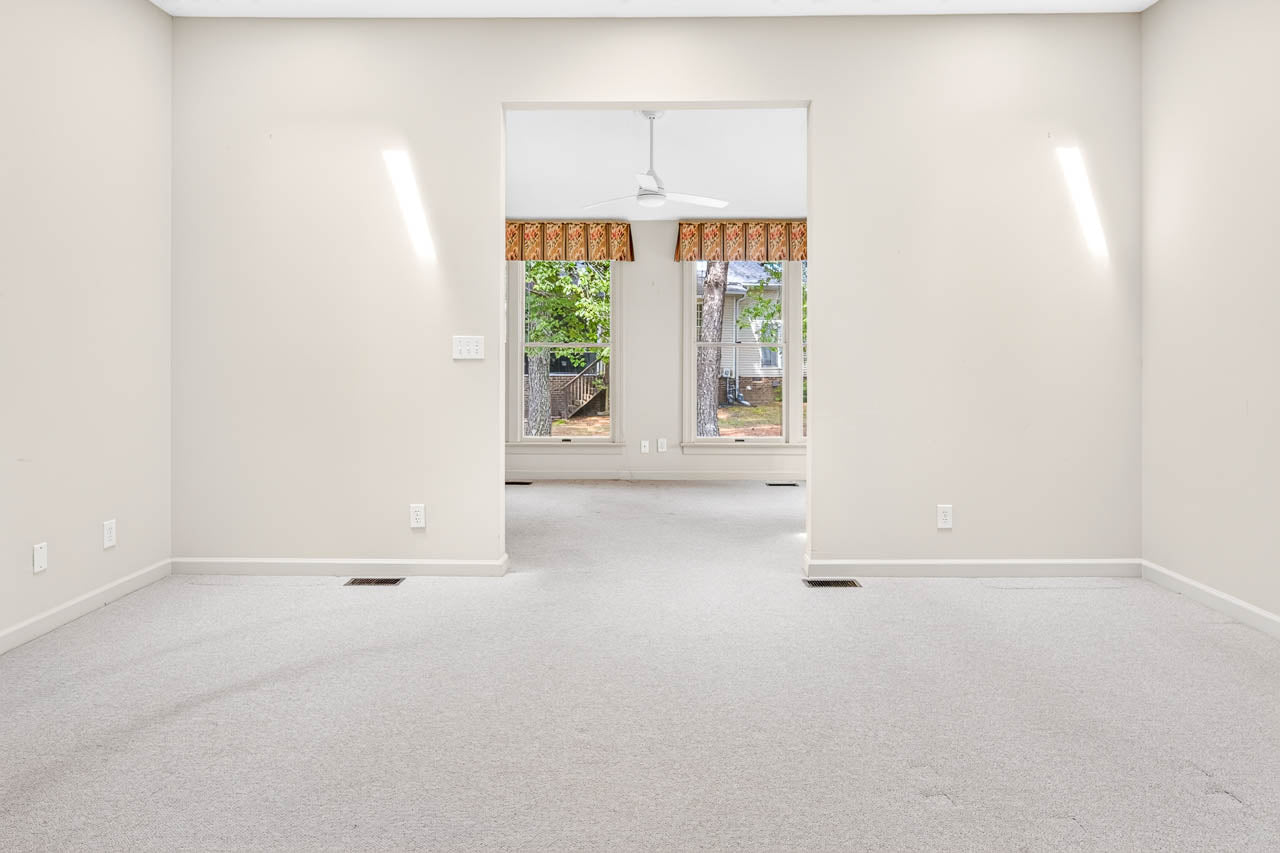 Empty room with light beige walls and carpet, leading to another room with large windows, patterned valances, a ceiling fan, and a view of trees outside. The space is brightly lit with natural light.