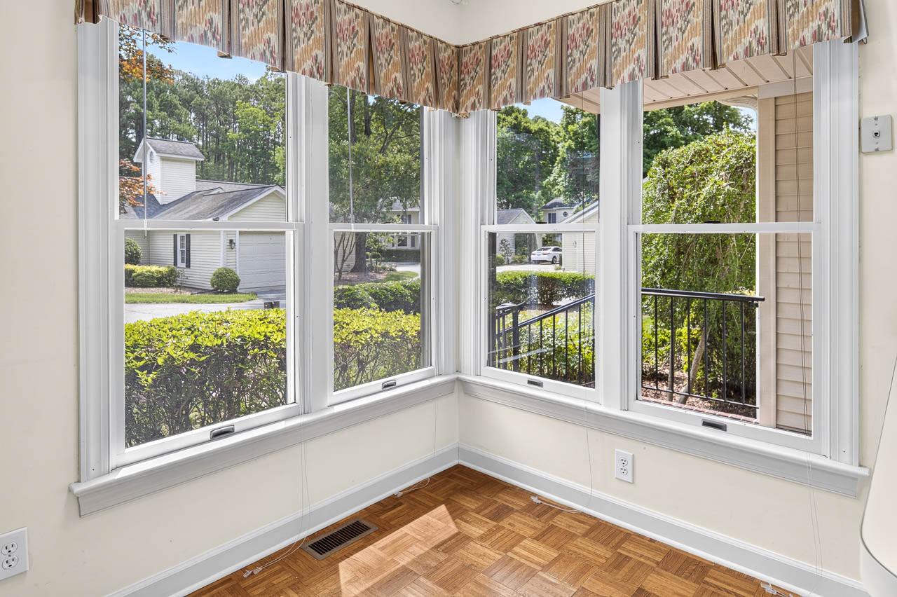 Corner of a sunlit room with large windows, wooden parquet floor, and patterned valance. The view shows a residential street, greenery, and houses outside.