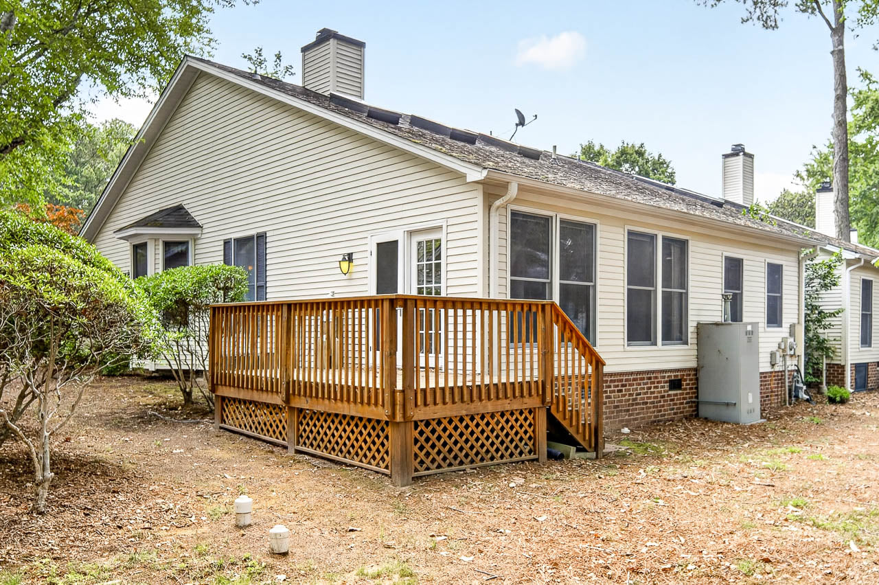 Single-story beige house with white trim, large windows, and a small wooden deck in the backyard. The yard has mulch, some shrubs, and trees, with utility boxes near the house.