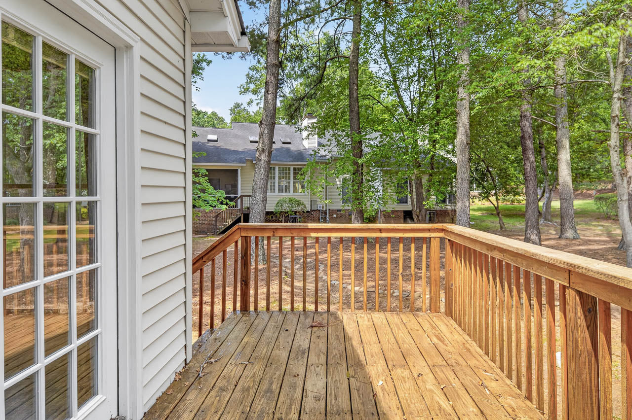 A wooden deck with railings extends from a house with white siding and glass doors, overlooking a wooded backyard and neighboring homes among tall trees.