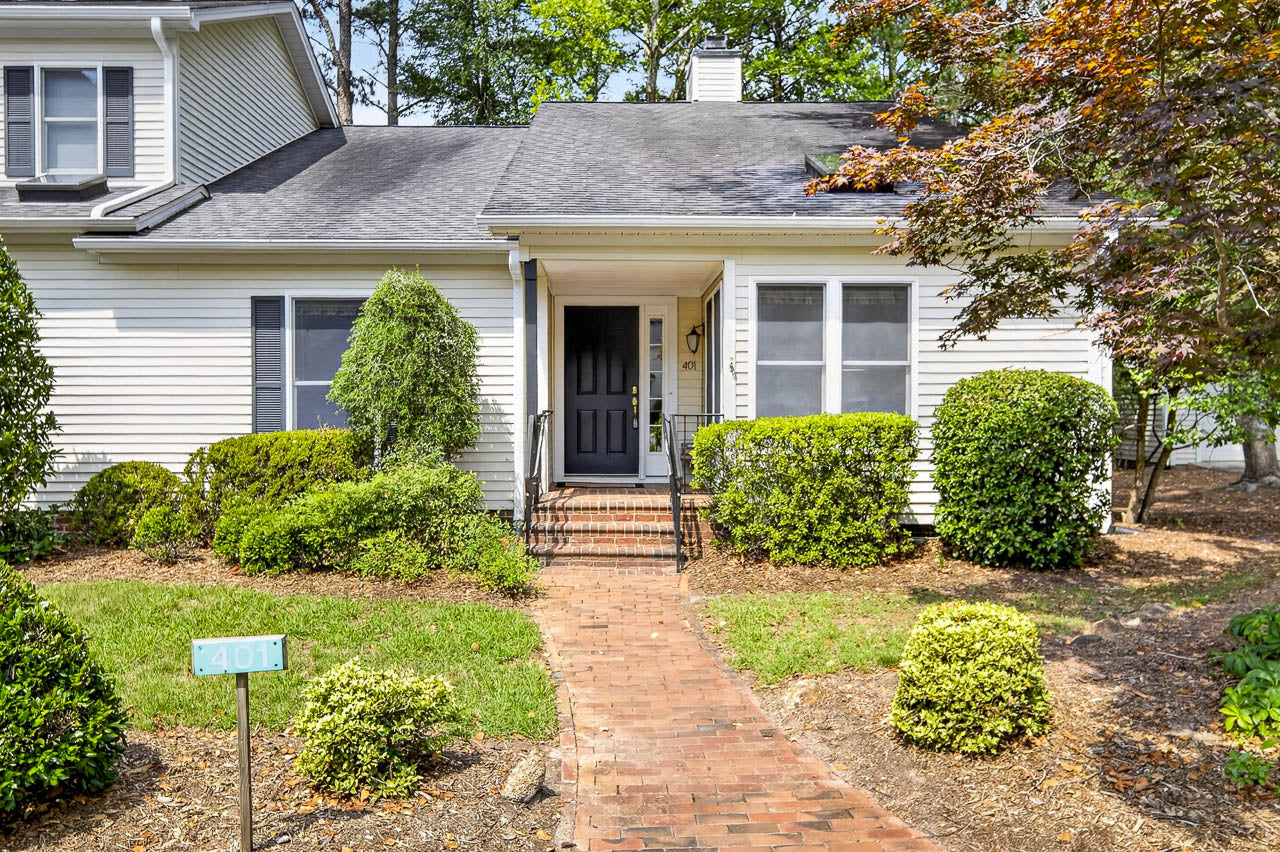 A white house with black shutters, a black front door, and brick steps leading up to a covered porch, surrounded by well-maintained green bushes and trees, with a house number sign "401" in the front yard.