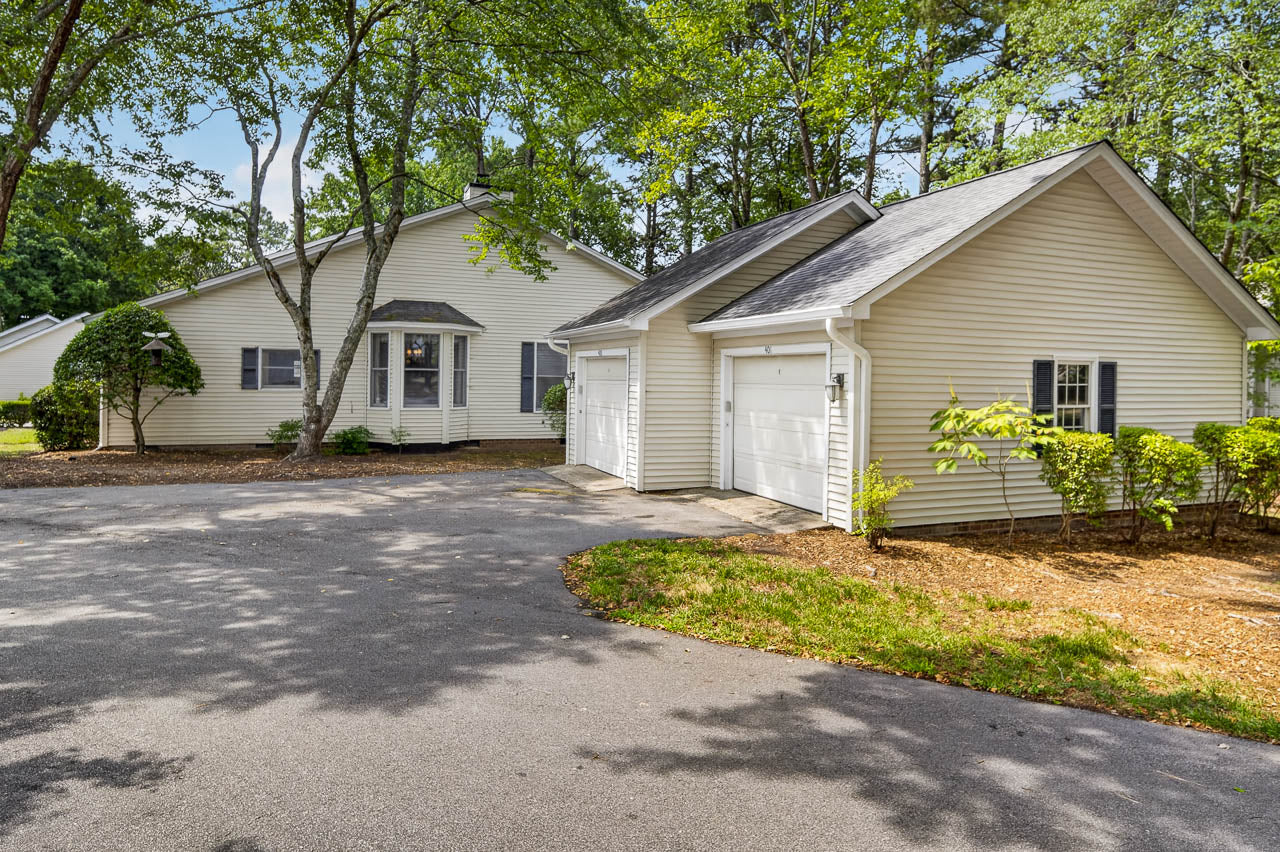 Single-story beige house with a detached two-car garage, surrounded by tall trees and greenery. The driveway curves in front, and the yard is landscaped with bushes and mulch.