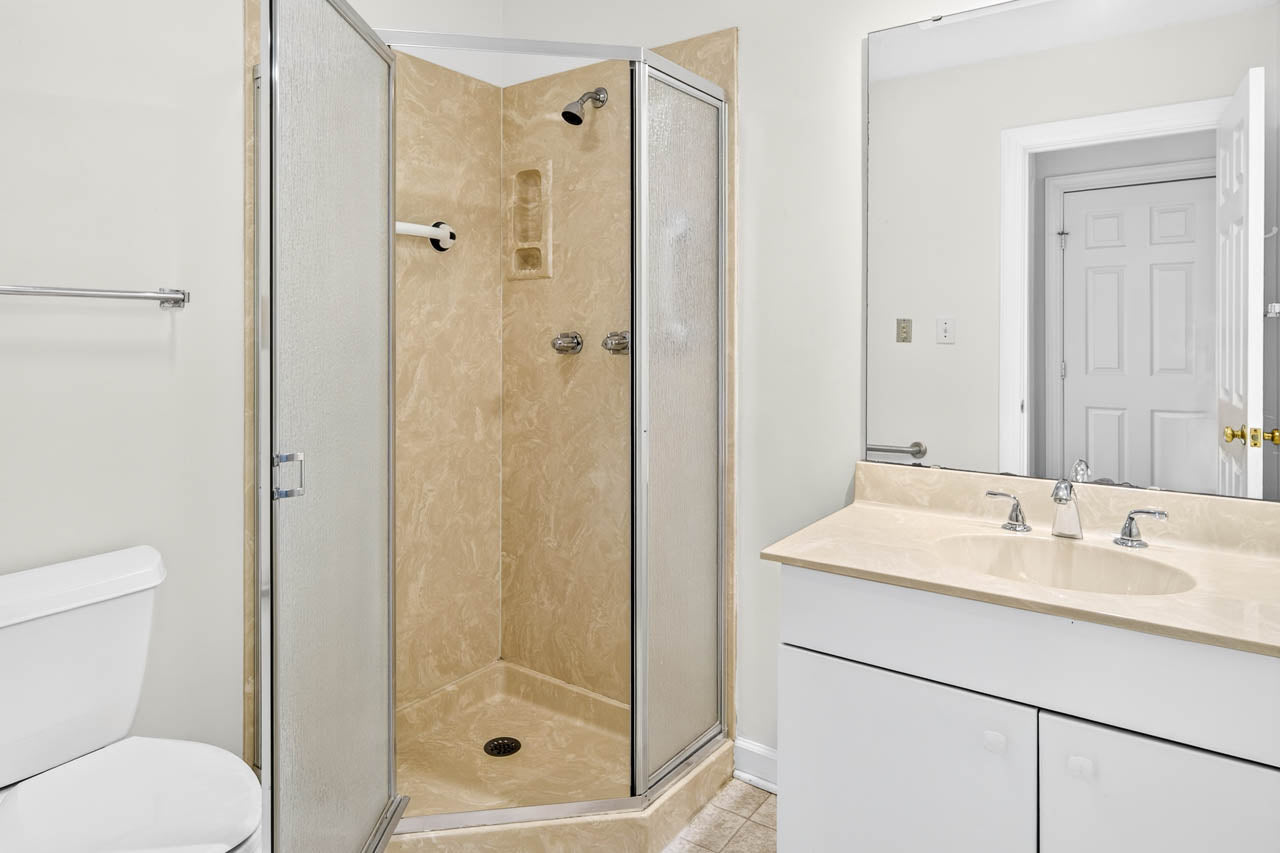 A bathroom with a corner shower featuring frosted glass doors, a white toilet, and a white vanity with a beige countertop and sink, reflected in a large mirror.