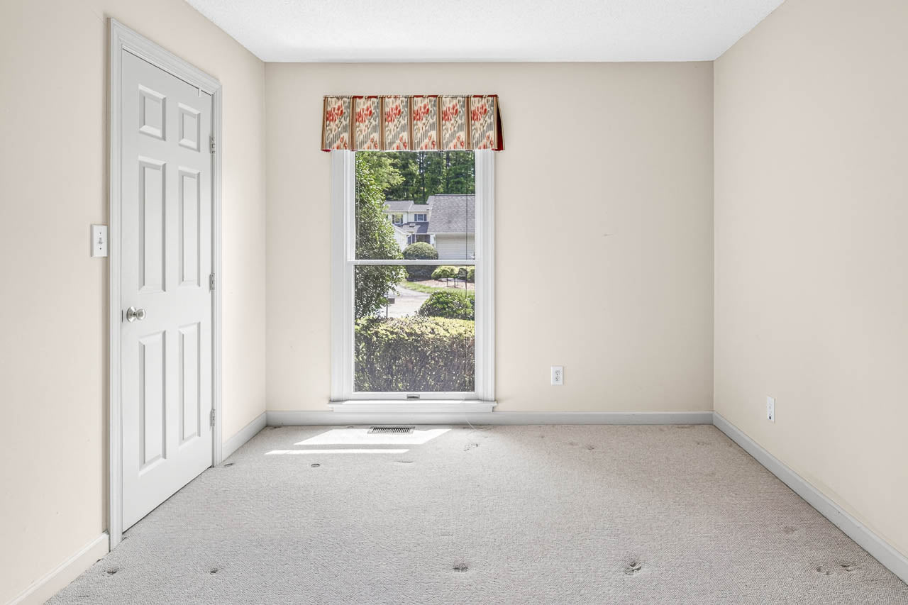 Empty beige room with carpeted floor, white door on the left, large window with floral valance in the center, and view of bushes and a house outside. The walls are plain and there is no furniture.