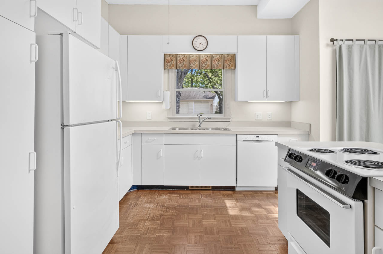 A bright, clean kitchen with white cabinets, white appliances, a window above the sink, a stove, a fridge, and wood-patterned flooring. A small clock and a patterned valance are above the window.