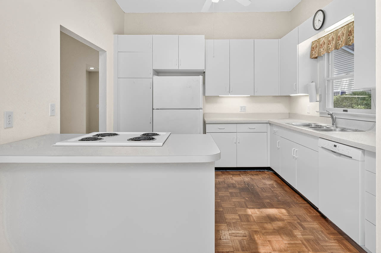 Bright kitchen with white cabinets and appliances, a built-in stovetop on an island, double sink under a window with a valance, dishwasher, and wood flooring. A wall clock is above the window.