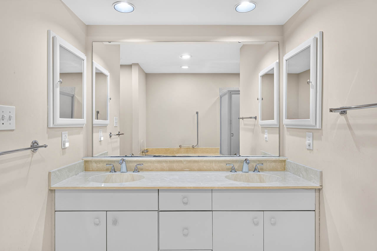 A bathroom with a double-sink vanity, white cabinets, and a large mirror. Two wall-mounted medicine cabinets with mirrors are on each side. The walls are beige, and the lighting is recessed ceiling lights.