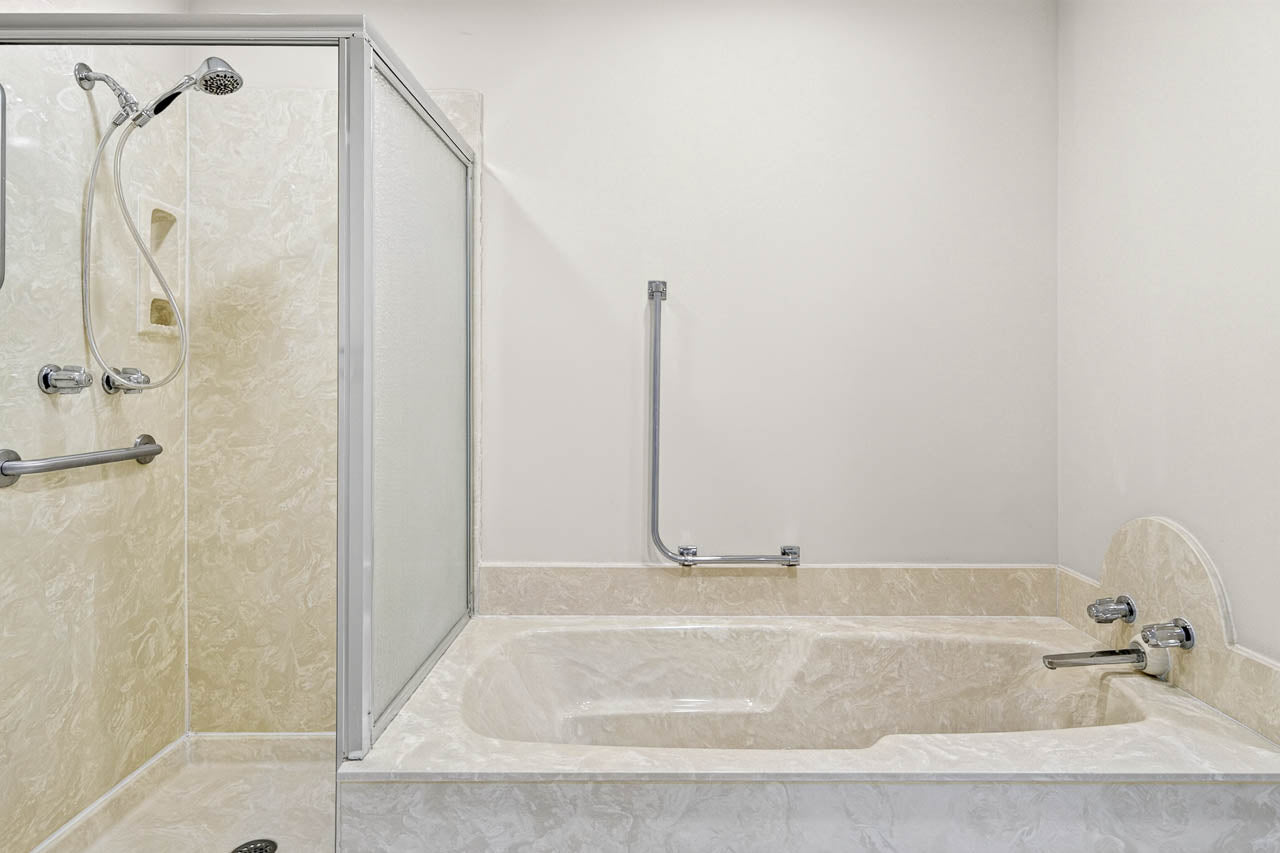 A bathroom with a beige marble-style bathtub and a separate shower enclosure. The bathtub has two faucets and a grab bar on the wall. The shower has a handheld showerhead and grab bars for safety.