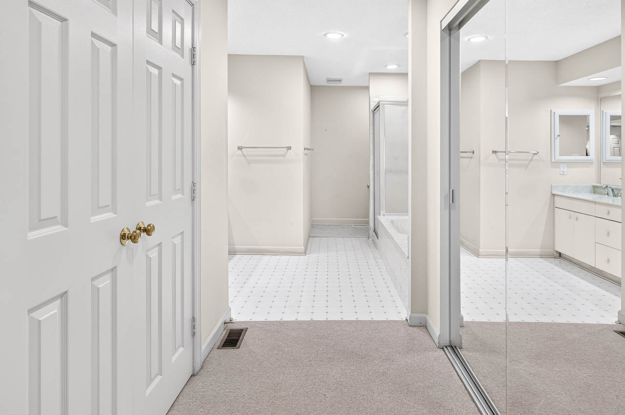 Spacious bathroom with beige walls, white tile flooring, double sinks, a large vanity mirror, a bathtub with a glass shower door, towel racks, and mirrored closet doors. Carpeted area leads into the bathroom.