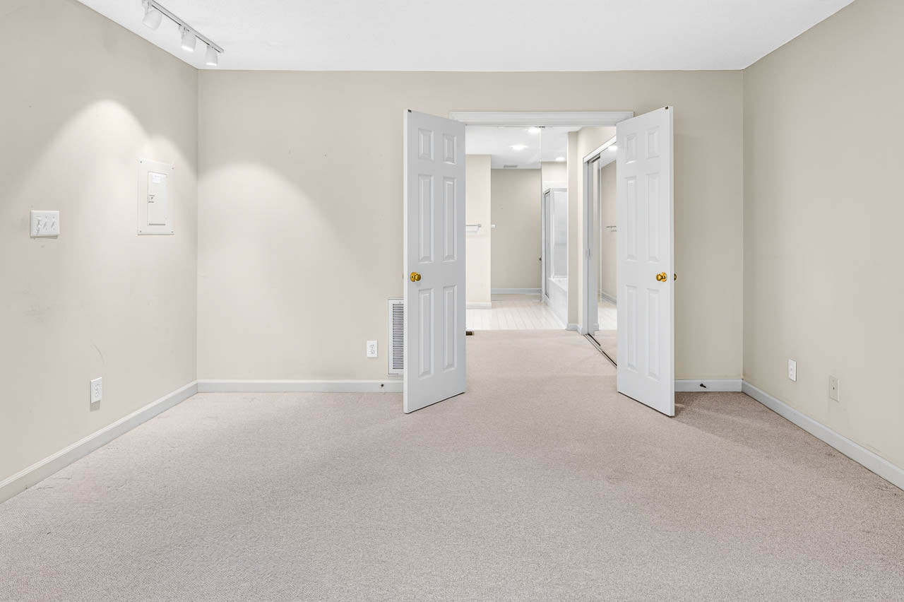 Spacious, empty room with beige carpet and walls, double white doors open to a bright hallway, and ceiling lights illuminating the area.