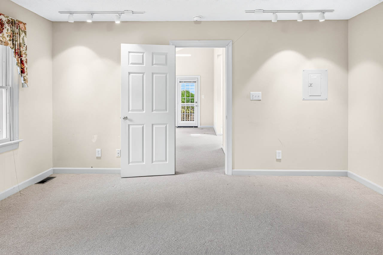 A bright, empty room with beige walls and carpet, an open white door leading to another room with windows and natural light. Track lighting is on the ceiling, and a window with floral curtains is on the left.