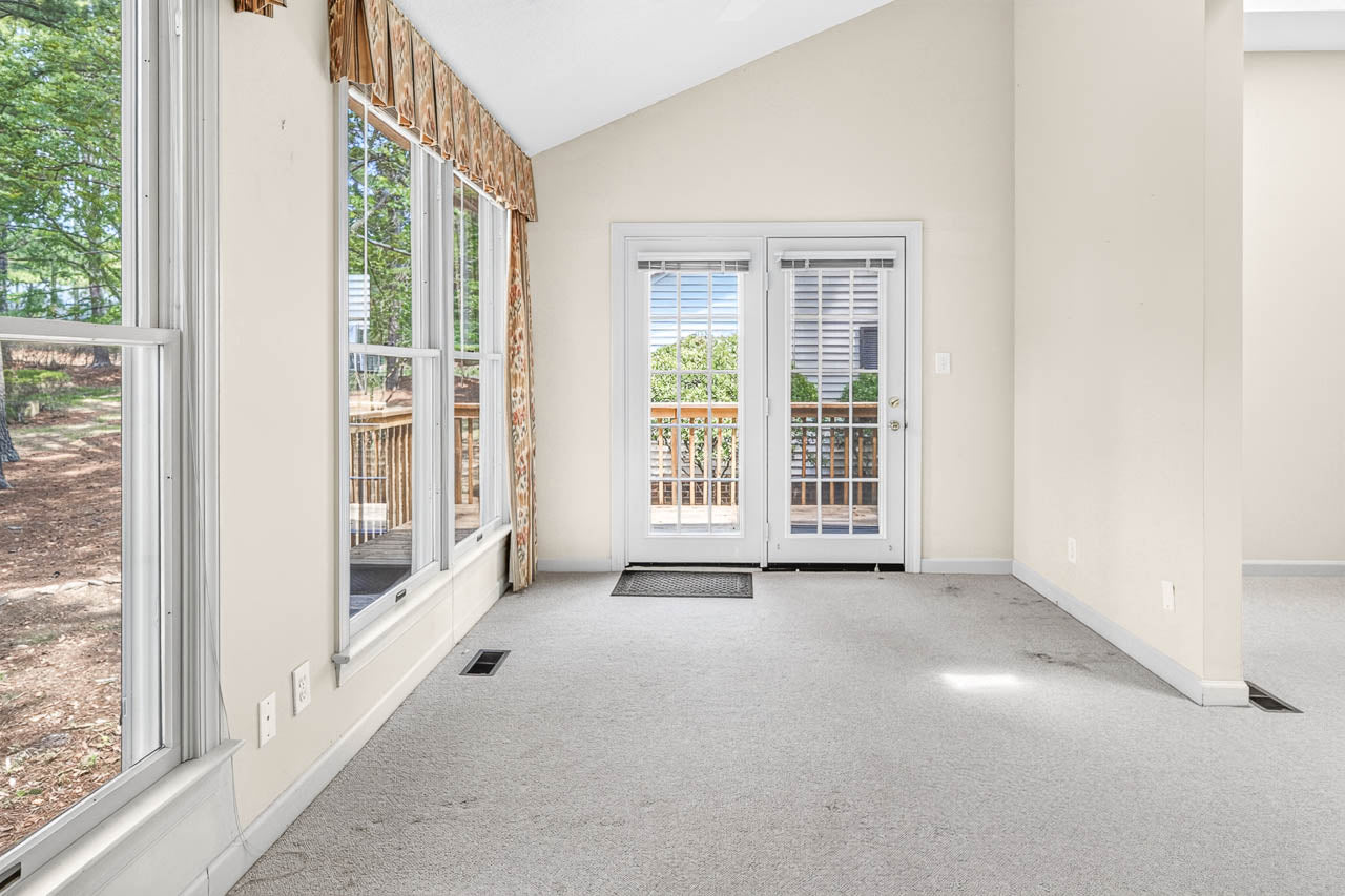 Bright, empty room with light carpet, large windows, and French doors opening to a wooden deck outside. Beige walls and a patterned valance above the windows add a warm touch. Natural light fills the space.