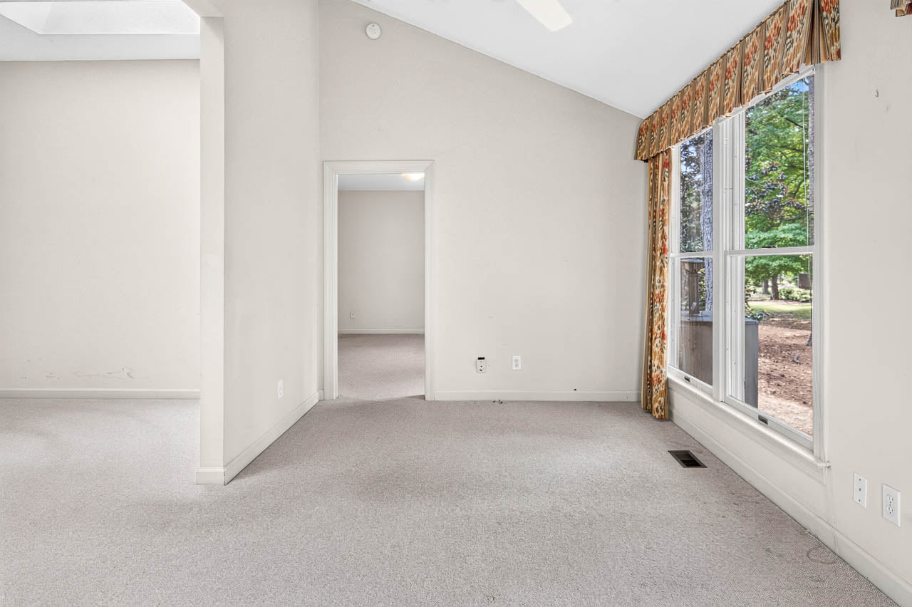 A bright, empty room with beige carpet and walls, large window with patterned curtains on the right, and a doorway leading to another room. Trees and greenery are visible outside the window.