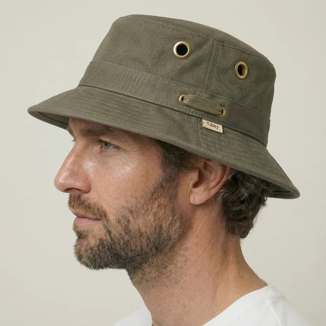 A person wearing the TILLEY - ICONIC T1 BUCKET HAT IN OLIVE with yellow eyelets and a white crew-neck shirt looks slightly to the side against a plain light background. The TILLEY hat offers UPF 50+ sun protection.