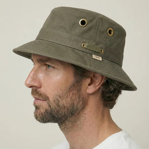 A person wearing the TILLEY - ICONIC T1 BUCKET HAT IN OLIVE with yellow eyelets and a white crew-neck shirt looks slightly to the side against a plain light background. The TILLEY hat offers UPF 50+ sun protection.